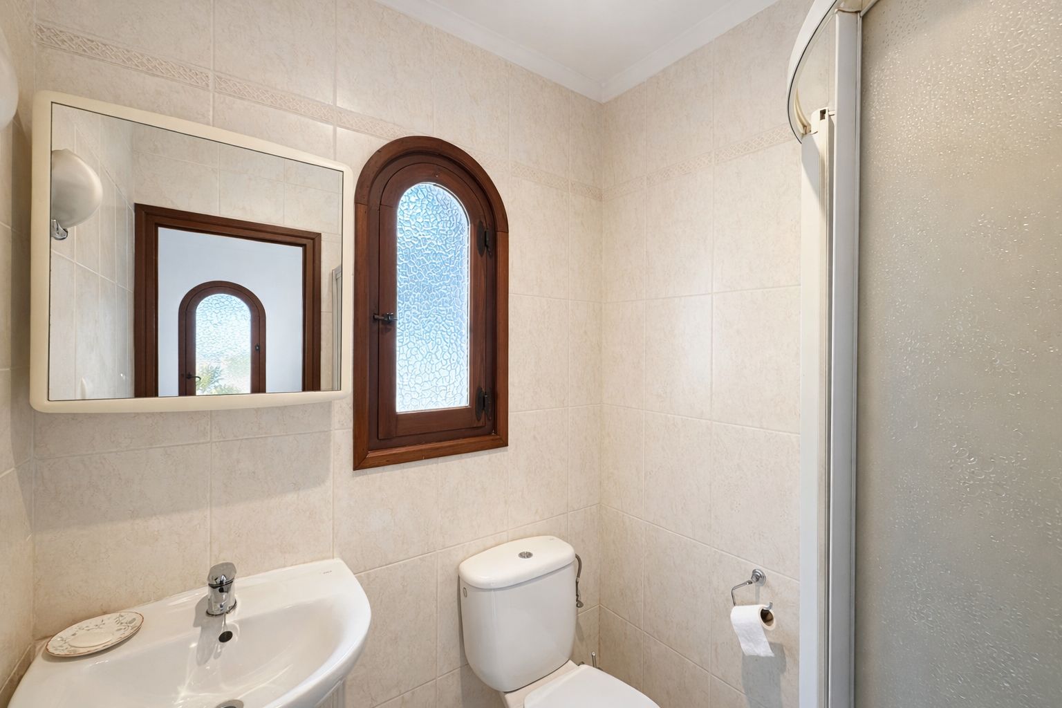 Badezimmer mit beigefarbenen Wandfliesen, einem Rundbogenfenster aus Holz mit Strukturglas und weißem Keramikwaschbecken.