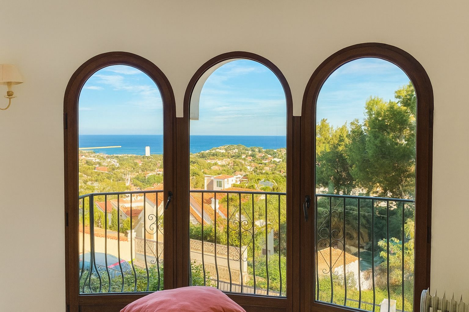 Drei Rundbogenfenster mit dunklen Holzrahmen bieten einen Panoramablick auf das Mittelmeer und die Küstenlandschaft, ergänzt durch ein schmiedeeisernes Geländer.
