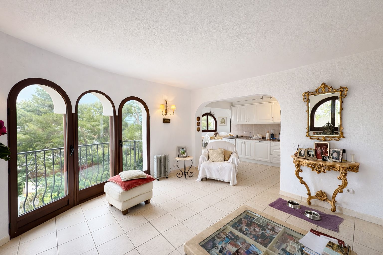 Helles Wohnzimmer mit drei Rundbogen-Fenstertüren zum Balkon, Blick ins Grüne, hellen Fliesenböden und einer verzierten goldenen Konsole.