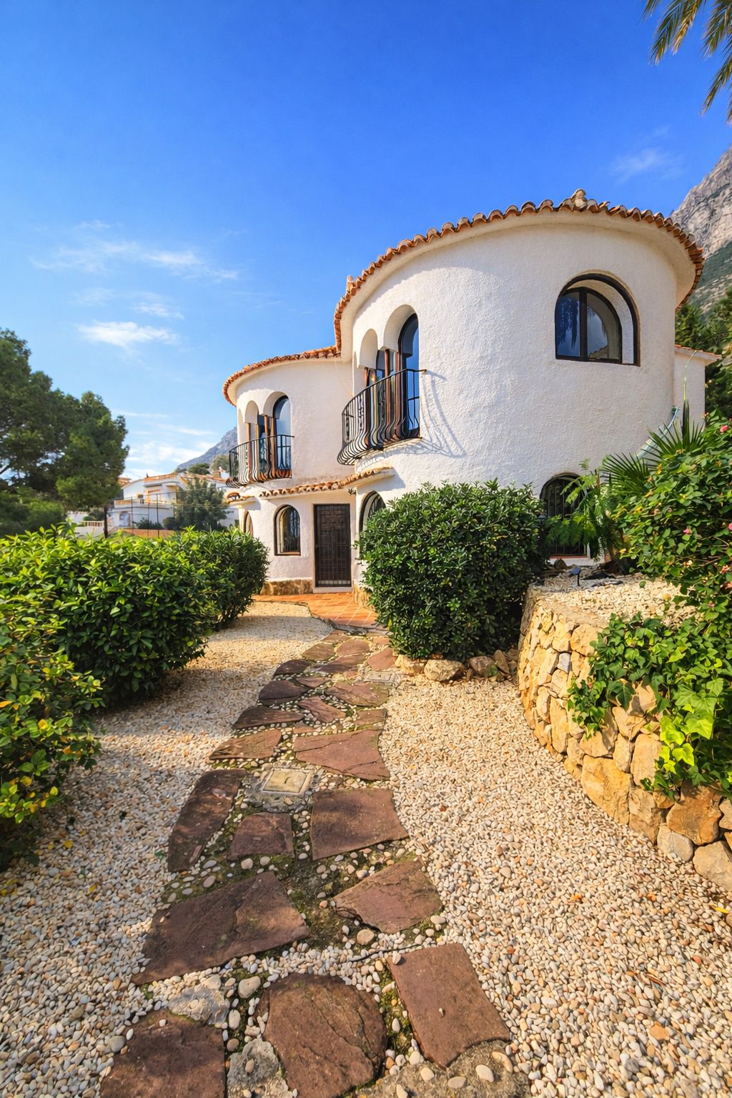 Mediterrane Villa mit geschwungenen weißen Wänden, Rundbogenfenstern und schmiedeeisernen Balkonen. Ein Natursteinpfad führt durch den Kiesgarten mit Hecken.
