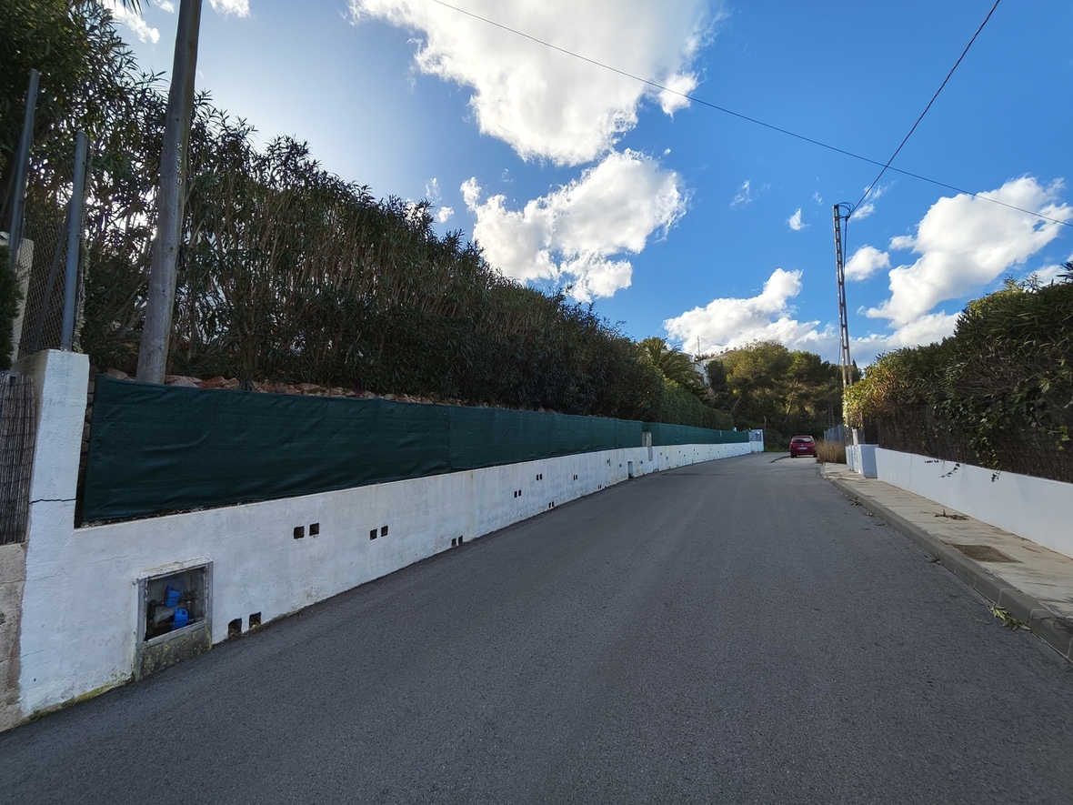 Ansicht der Zufahrtsstraße mit weißer Begrenzungsmauer, grünem Sichtschutznetz und hohen Hecken unter einem blauen, leicht bewölkten Himmel.