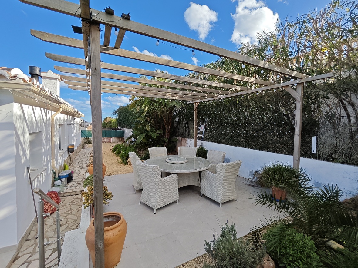 Außen-Essbereich mit rundem Glastisch und sechs weißen Korbstühlen unter einer Holzpergola mit Lichterketten, umgeben von mediterraner Bepflanzung.