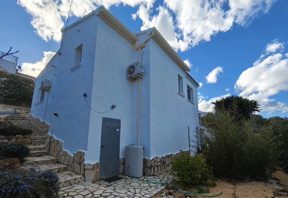 Außenansicht der Villa mit weiß verputzten Wänden, einem Natursteinsockel und installierten Klimageräten vor einem blauen, bewölkten Himmel.
