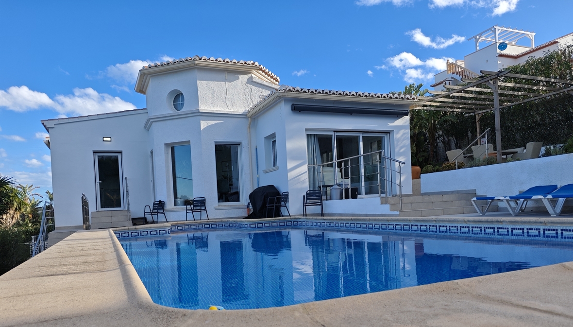 Außenansicht einer weißen mediterranen Villa mit privatem Pool, Sonnenterrasse mit blauen Liegen und einer Holzpergola unter klarem Himmel.