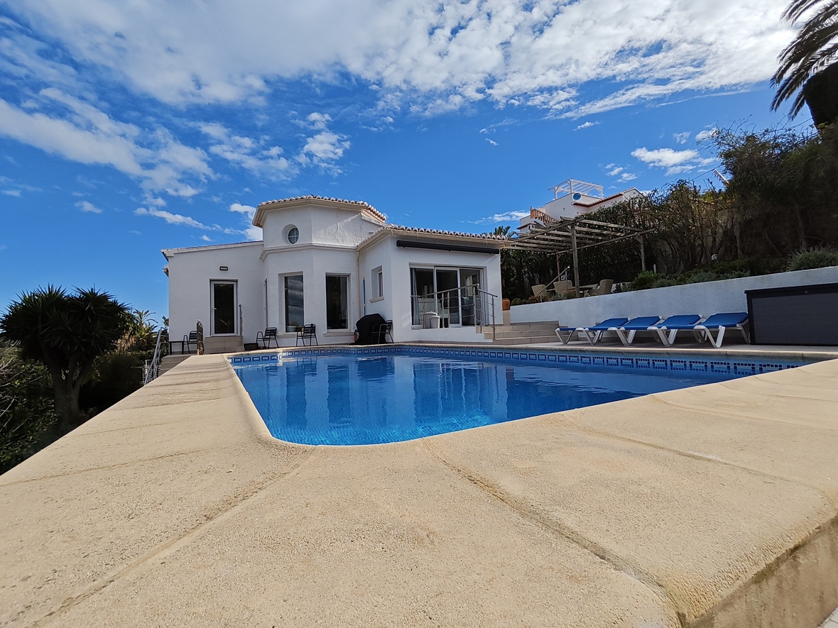 Außenansicht einer weißen Villa mit privatem Pool, Steinterrasse, blauen Sonnenliegen und mediterraner Architektur unter blauem Himmel.