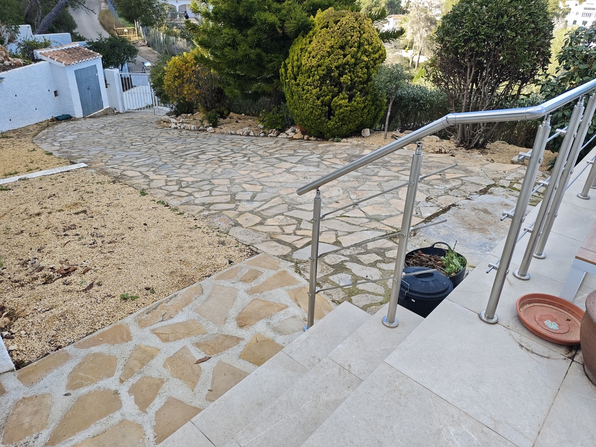 Außenansicht mit einem natursteingepflasterten Weg, umgeben von mediterraner Bepflanzung und einer Treppe mit Edelstahlgeländer.