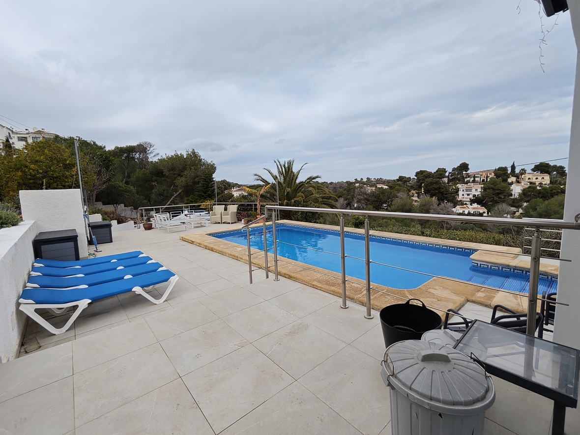 Erhöhte Terrasse mit rechteckigem Pool, Steineinfassung, blauen Sonnenliegen und Edelstahlgeländer mit Blick auf ein bewaldetes Wohngebiet.