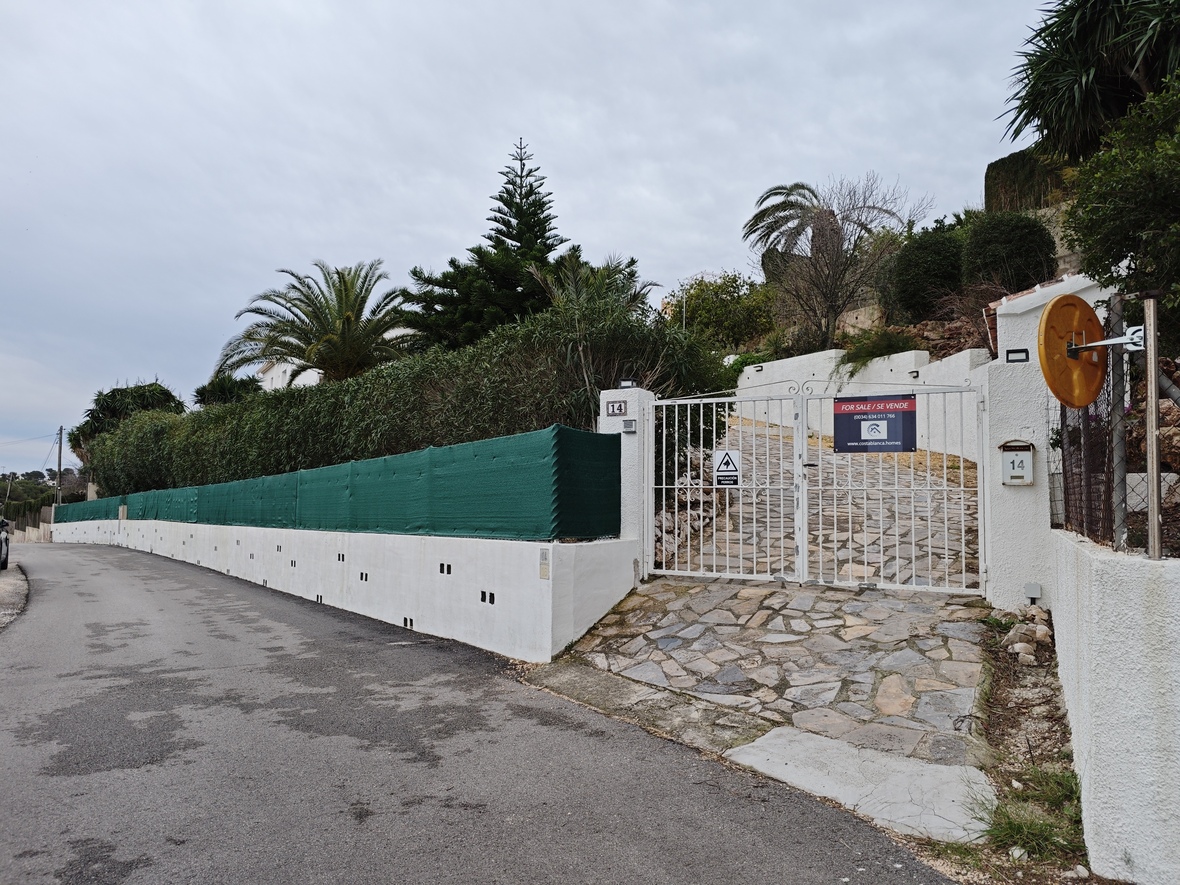 Haupteingang mit weißem Metalltor, gepflasterter Steinfahrt und einer Umfassungsmauer mit Sichtschutz und mediterraner Bepflanzung.