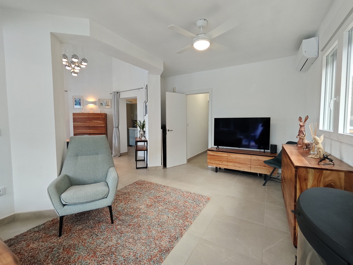 Heller Wohnbereich mit Polstersessel, TV-Möbeln aus Naturholz und Flachbildfernseher. Ausgestattet mit Deckenventilator, Klimaanlage und hellen Fliesenböden.