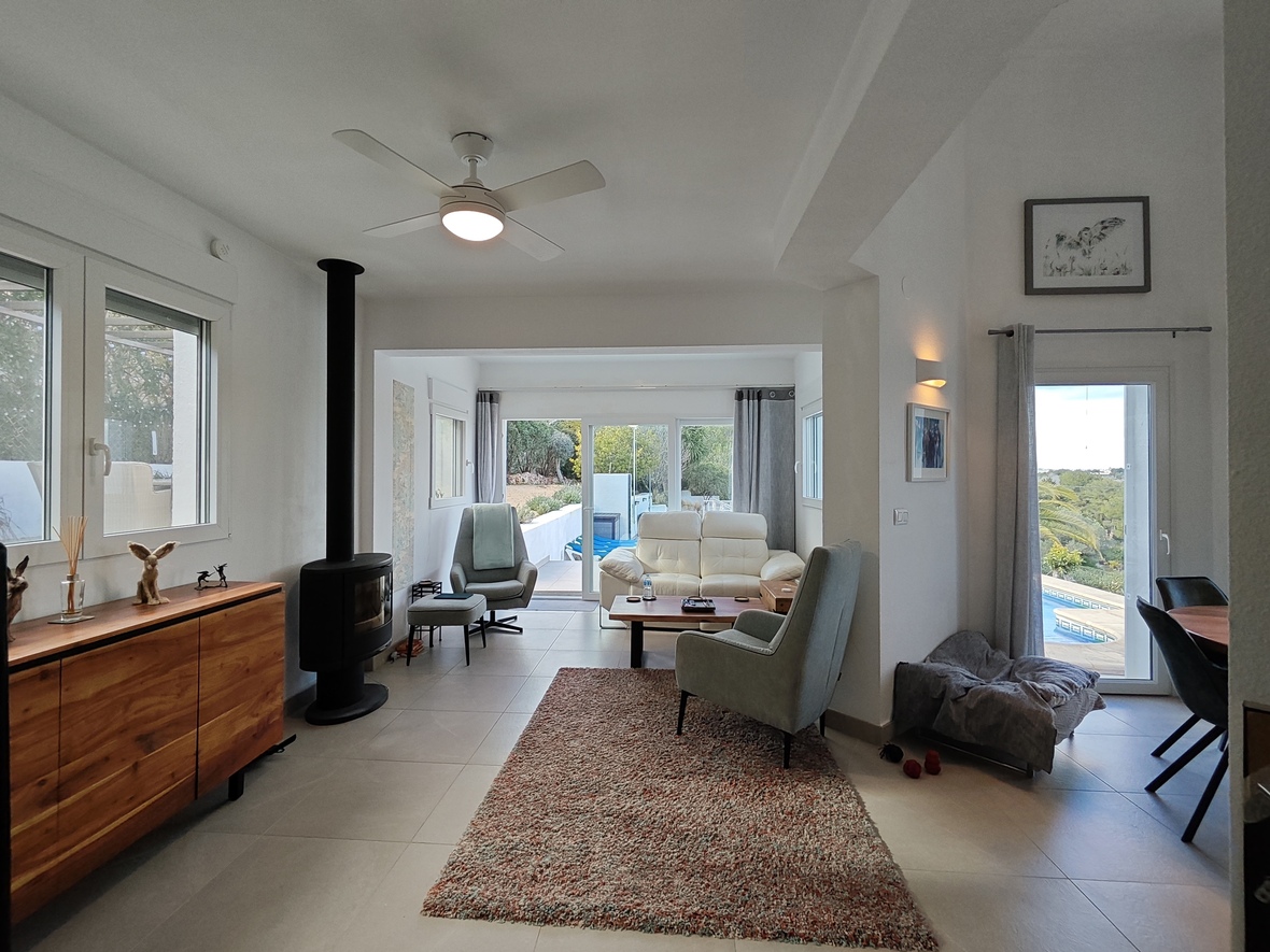 Helles Wohnzimmer mit schwarzem Kaminofen, Sideboard aus Holz und Deckenventilator. Große Glastüren führen direkt zur Terrasse und zum Poolbereich.
