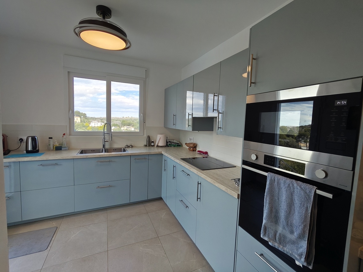 Moderne Küche mit hellblauen Fronten, Arbeitsplatte in Holzoptik und integrierten Samsung-Geräten. Ein großes Fenster bietet Ausblick auf die umliegende Landschaft.