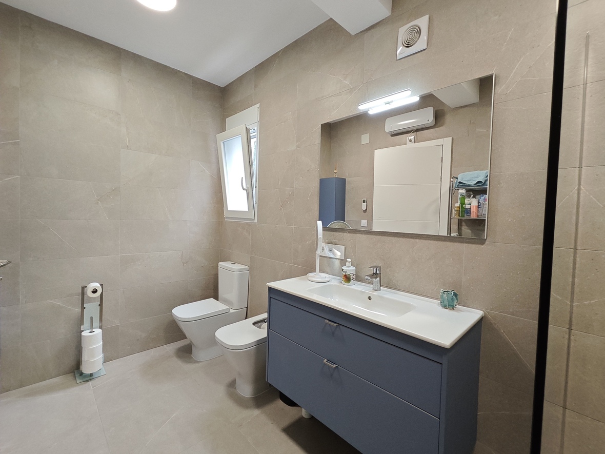 Modernes Badezimmer mit beigen Steineffektfliesen, blauem Hängewaschbecken-Unterschrank und passendem Bidet und WC. Ein großer Spiegel mit LED-Leiste sorgt für Licht.