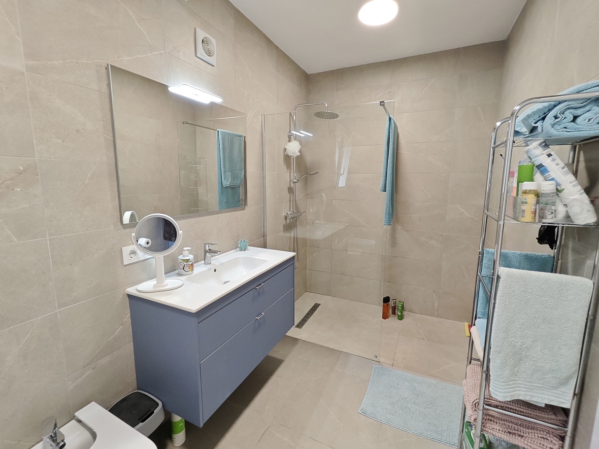 Modernes Badezimmer mit blauem Hängeschrank, begehbarer Dusche mit Glastrennwand und sandfarbenen großformatigen Fliesen.