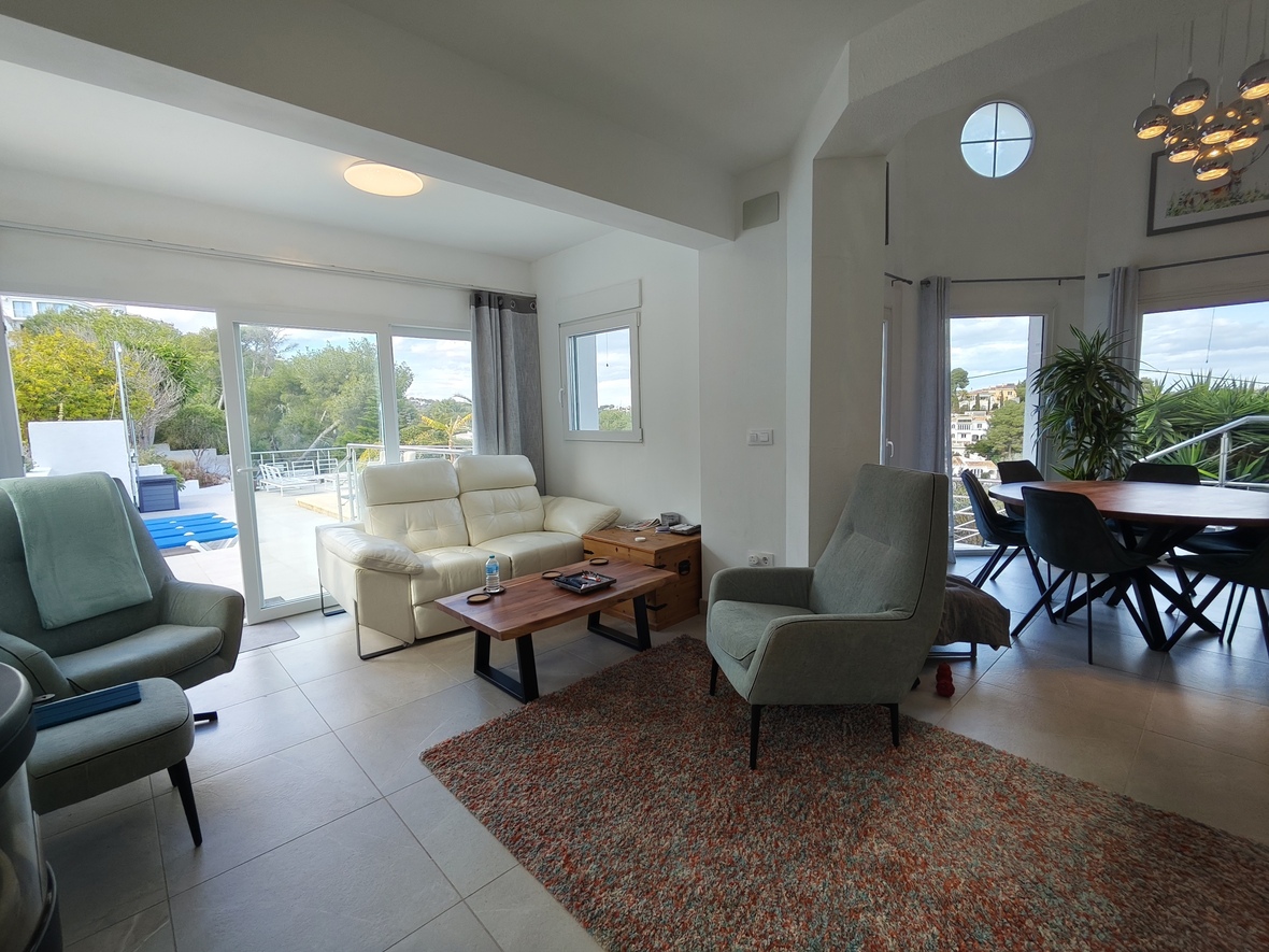 Offener Wohnbereich mit weißem Ledersofa, hohen Decken und großen Glasschiebetüren, die direkt auf die Terrasse mit Pool und Gartenblick führen.