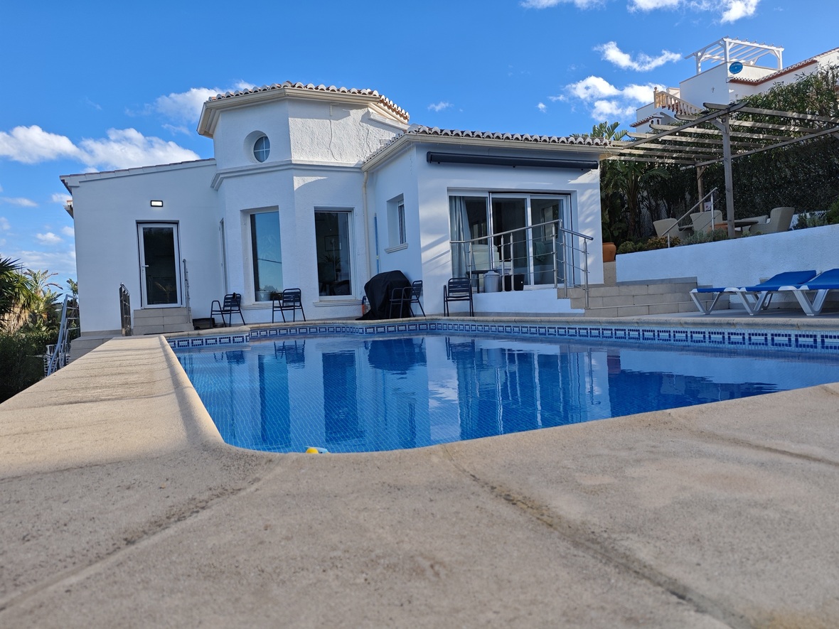 Privater Pool mit blauen Mosaikfliesen und Steinterrasse vor einer weißen mediterranen Villa mit markantem achteckigem Turm und großen Glasschiebetüren.