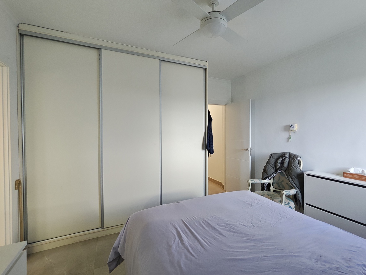 Schlafzimmer mit einem großen dreitürigen Schiebetürenschrank in Weiß, einem Deckenventilator und einem klassischen Sessel neben der Kommode.