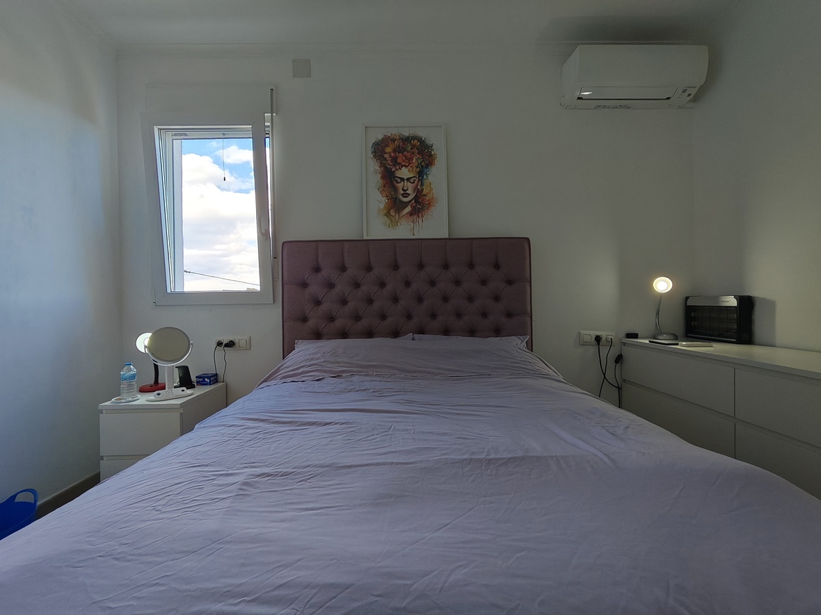 Schlafzimmer mit mauvefarbenem, gepolstertem Kopfteil, weißen minimalistischen Möbeln und Klimaanlage. Ein dekoratives Porträt hängt über dem Bett am Fenster.