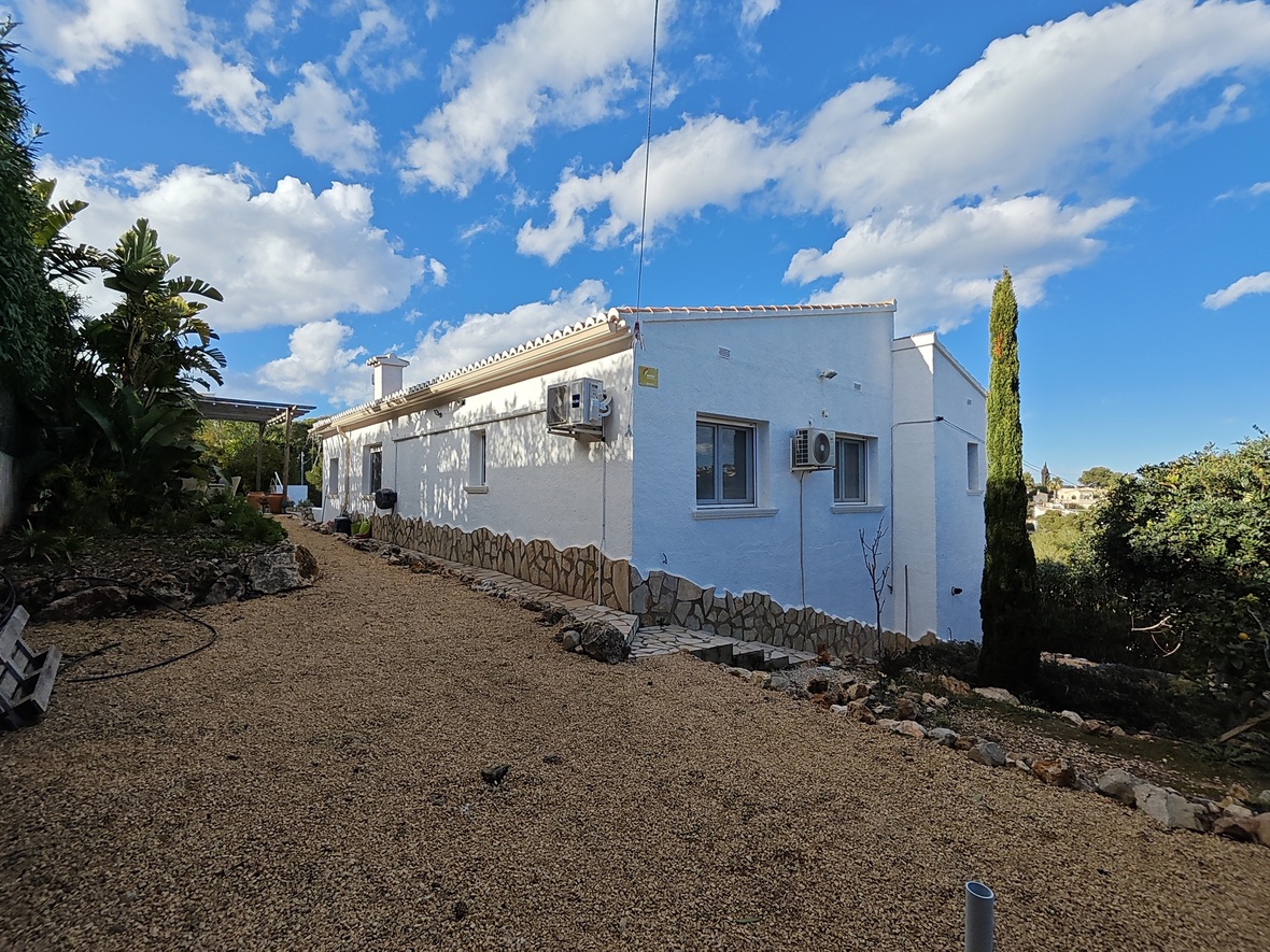 Seitenansicht einer weißen Villa mit Natursteinsockel, Kiesweg und wandmontierten Klimaanlagen, umgeben von mediterraner Bepflanzung und einer Zypresse.