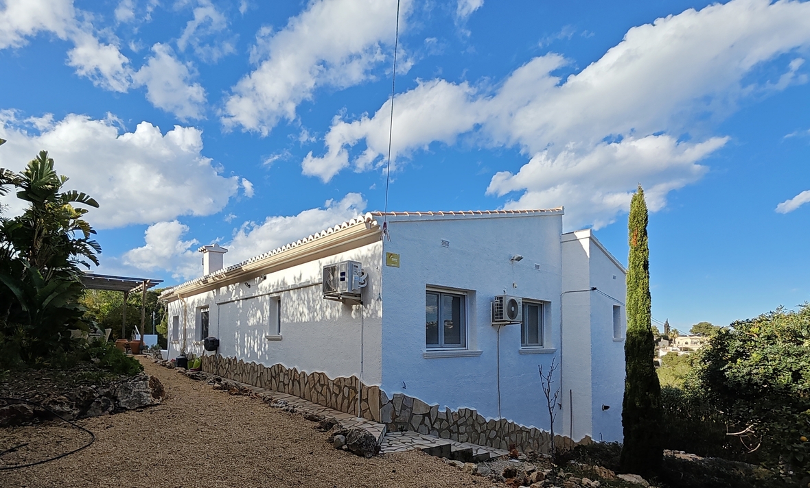 Weiße Außenfassade mit Natursteinsockel, Kiesweg und mediterraner Bepflanzung unter blauem Himmel.
