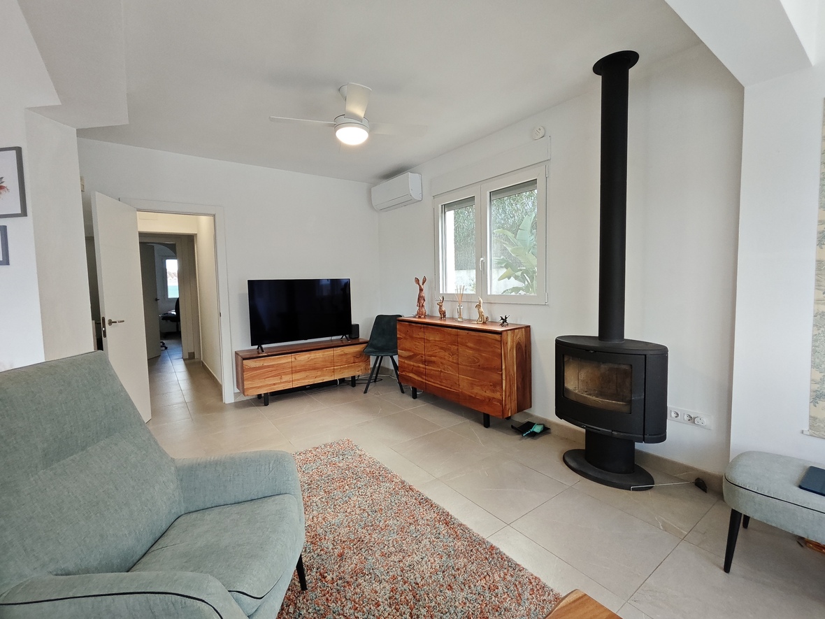 Wohnbereich mit modernem Kaminofen, Sideboards aus Massivholz und hellen Fliesenböden. Ausgestattet mit Klimaanlage, Deckenventilator und Blick ins Grüne.