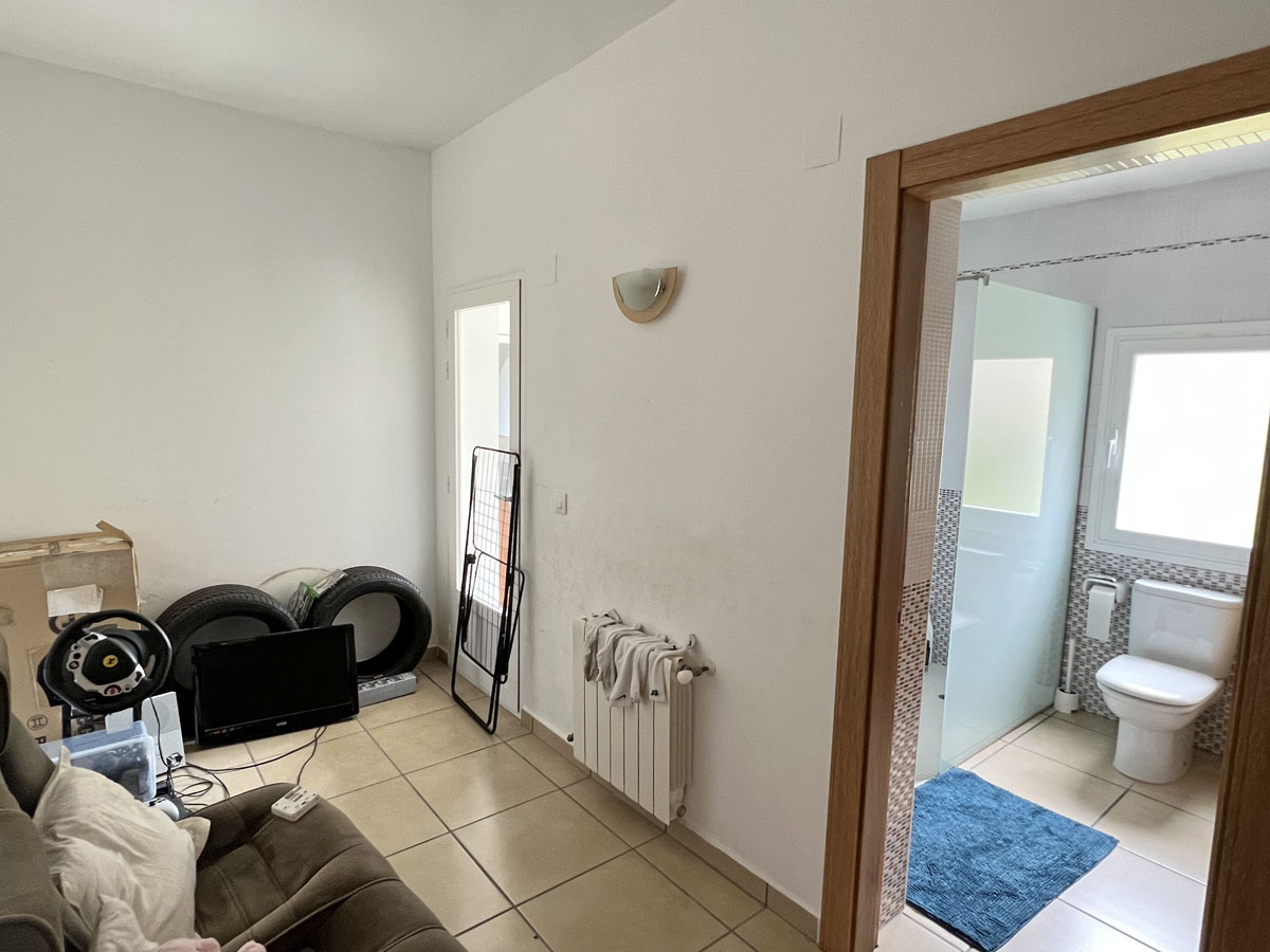Funktionaler Raum mit Fliesenboden und Heizkörper, verbunden mit einem Badezimmer mit begehbarer Dusche, Glastrennwand und Tageslicht.