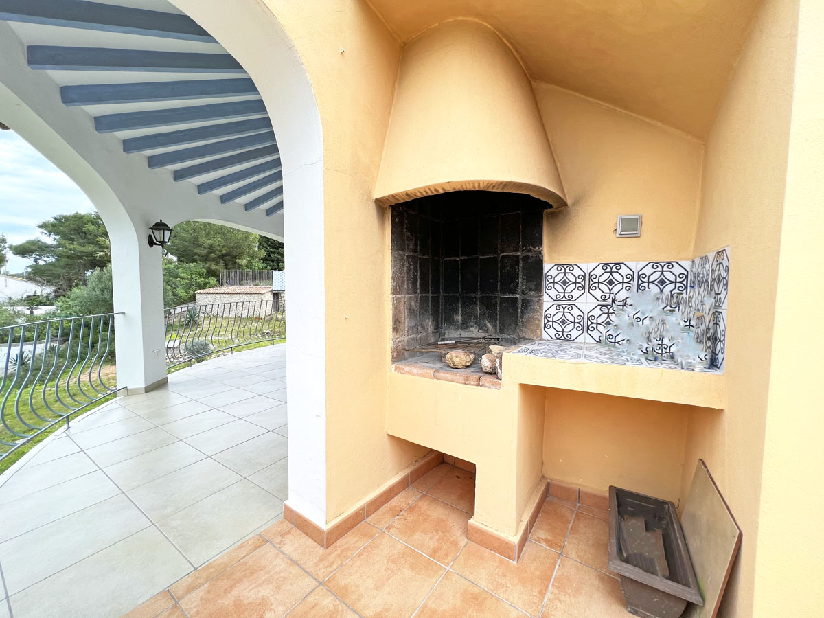 Gemauerter Außengrill mit konischer Haube und dekorativer Kachel-Arbeitsplatte auf einer überdachten Terrasse mit Rundbögen und blauen Deckenbalken.