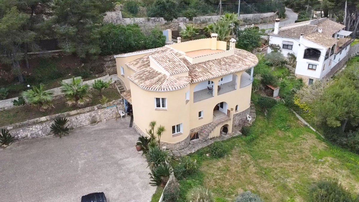 Luftaufnahme der mediterranen Villa mit geschwungener Fassade, Terrakotta-Ziegeldach und einer großen gepflasterten Einfahrt inmitten eines begrünten Grundstücks.