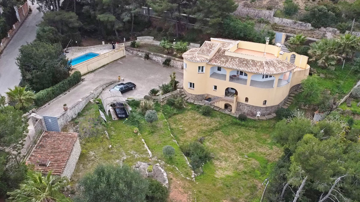 Luftaufnahme der Villa mit geschwungener Fassade und Steinsockel, großzügiger Parkfläche, privatem Pool und terrassiertem Garten inmitten mediterraner Vegetation.