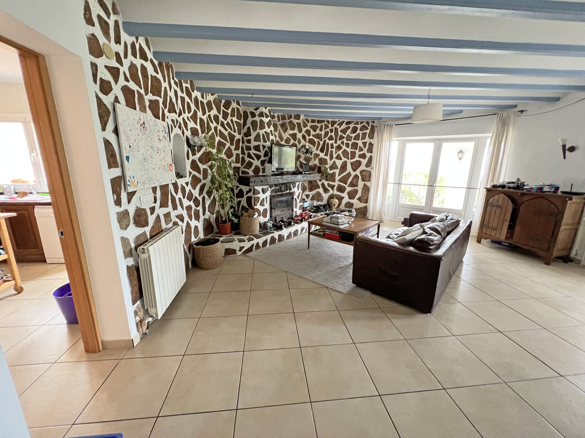 Rustikales Wohnzimmer mit Natursteinwand, integriertem Kamin, hellblauen Deckenbalken und Fliesenboden mit direktem Zugang zur Terrasse.