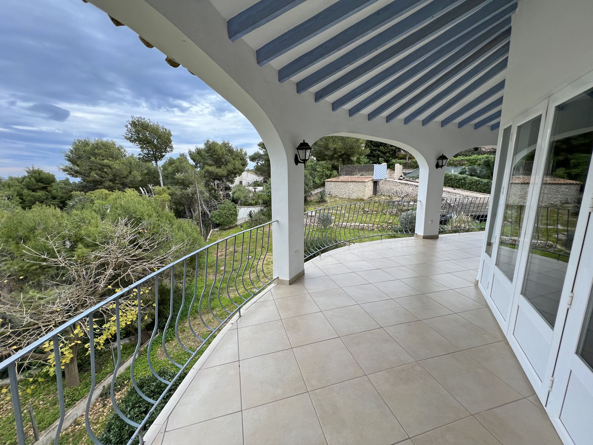 Überdachte Terrasse mit weißen Rundbögen, blauen Deckenbalken und Fliesenboden mit weitem Blick über den Garten bis zum Meer.