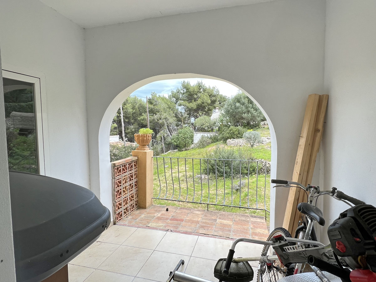 Überdachte Veranda mit Rundbogen und Blick auf den mediterranen Garten und Pinienwald, ausgestattet mit Fliesenboden und Metallgeländer.