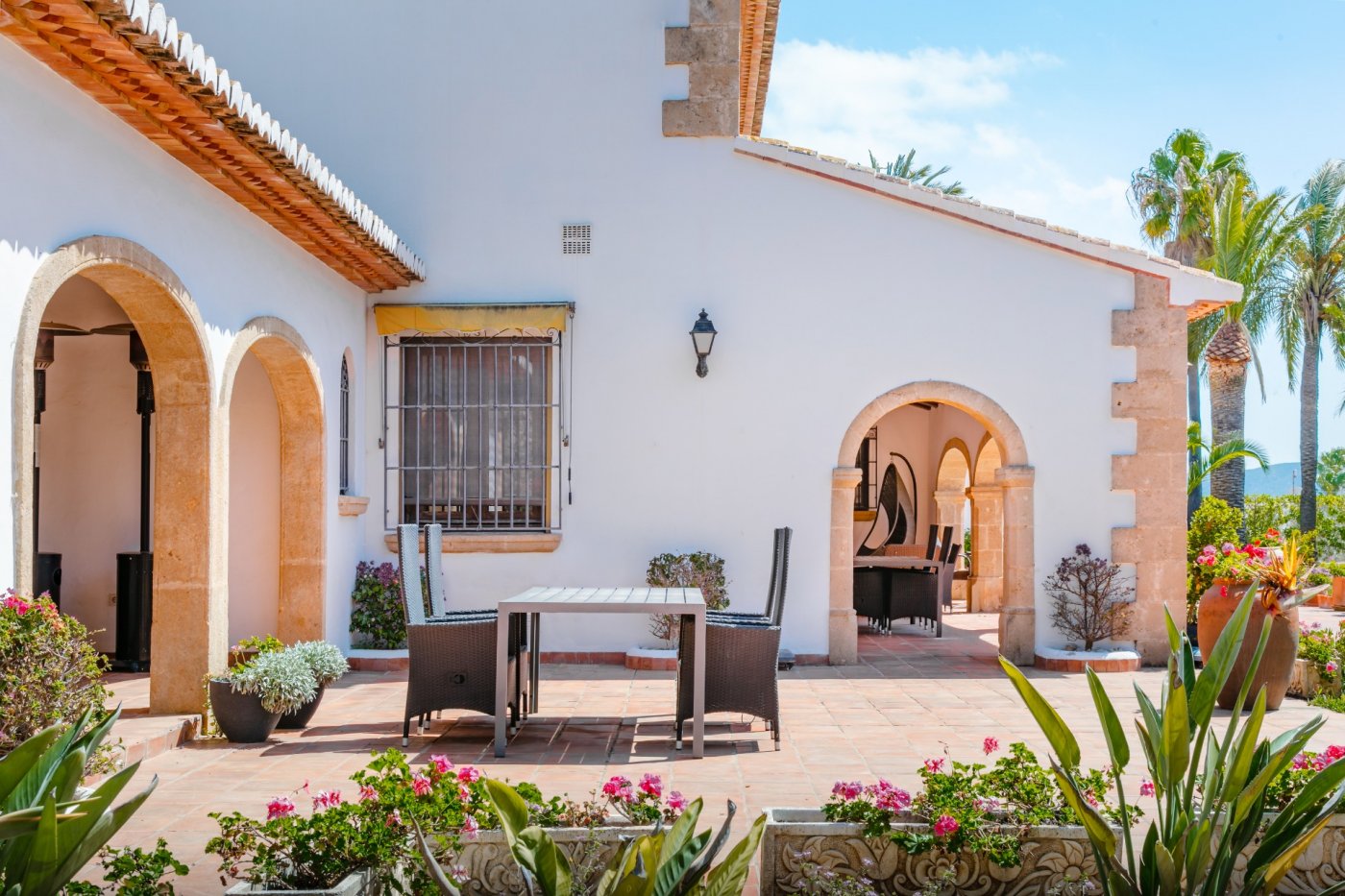 Außen-Terrasse mit traditionellen Tosca-Steinbögen, Terrakottaboden und Essbereich. Die weiße mediterrane Fassade wird von Palmen und Gartenpflanzen umgeben.