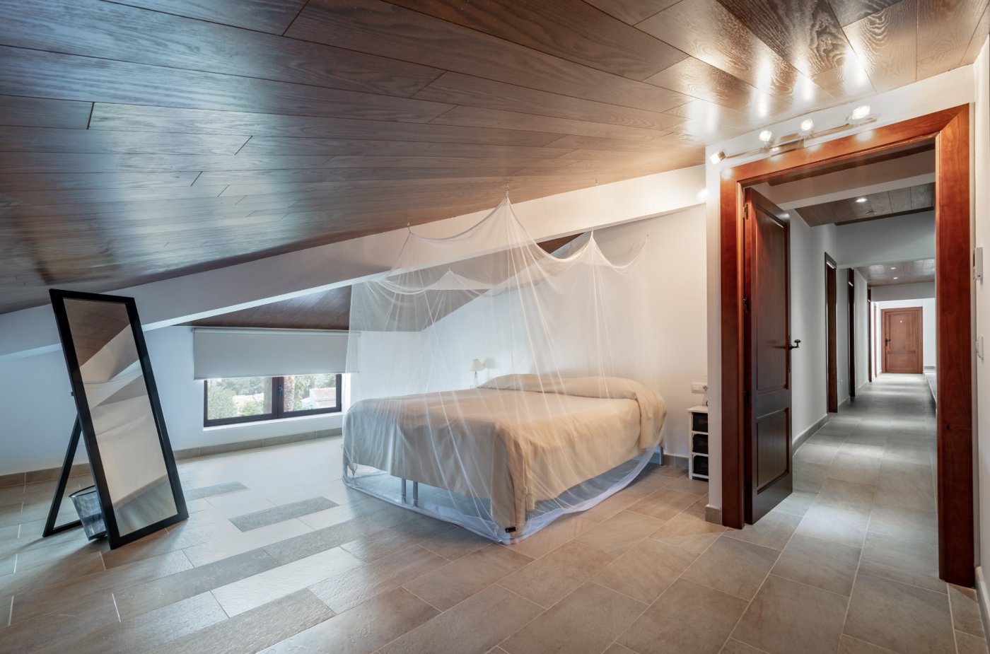 Dachgeschoss-Schlafzimmer mit dunkler Holzdecke, Steinfliesenboden und einem Bett mit Moskitonetz. Ein Flur mit Massivholz-Türrahmen verbindet die Räumlichkeiten der oberen Etage.