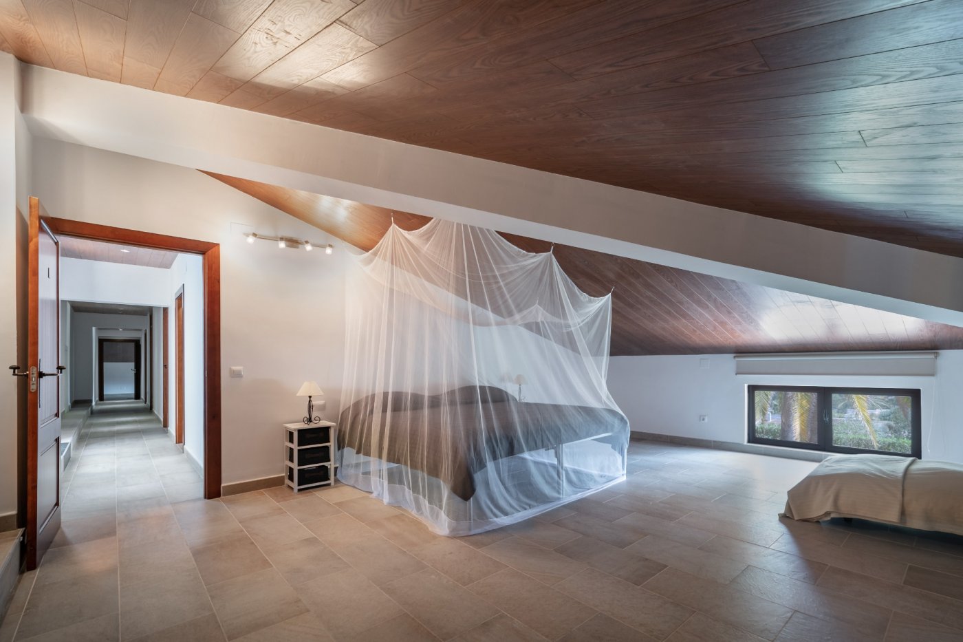 Dachgeschoss-Schlafzimmer mit Holzdecke, Fliesenboden und Bett mit Moskitonetz. Der Raum bietet Zugang zum Flur und einen Ausblick auf den mediterranen Garten.