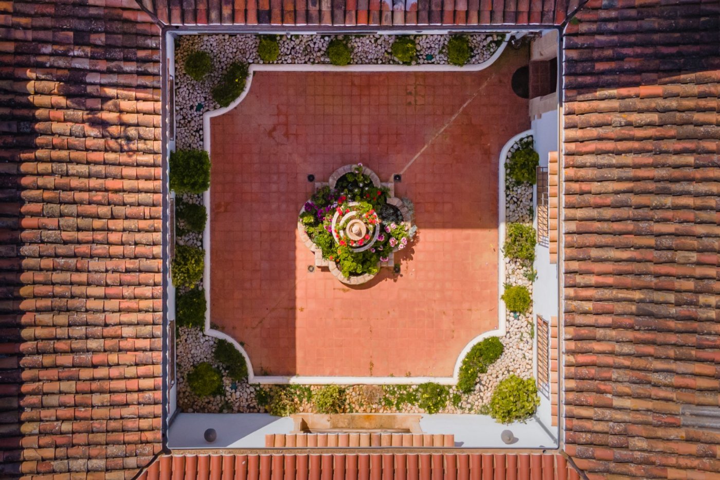 Draufsicht auf den zentralen Innenhof mit Terrakottaboden, einem Blumenbrunnen und Kiesbeeten, umrahmt von traditionellen mediterranen Ziegeldächern.