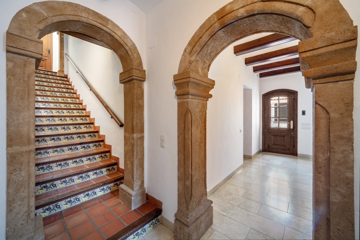 Eingangshalle mit traditionellen Rundbögen aus Tosca-Stein, einer Treppe mit dekorativen Keramikfliesen und einem Flur mit sichtbaren Holzbalken.