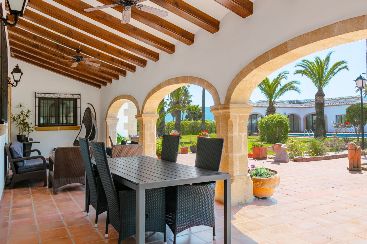 Großzügige überdachte Terrasse mit traditionellen Tosca-Steinbögen, Holzbalkendecken und Essbereich mit Blick auf den mediterranen Palmengarten.
