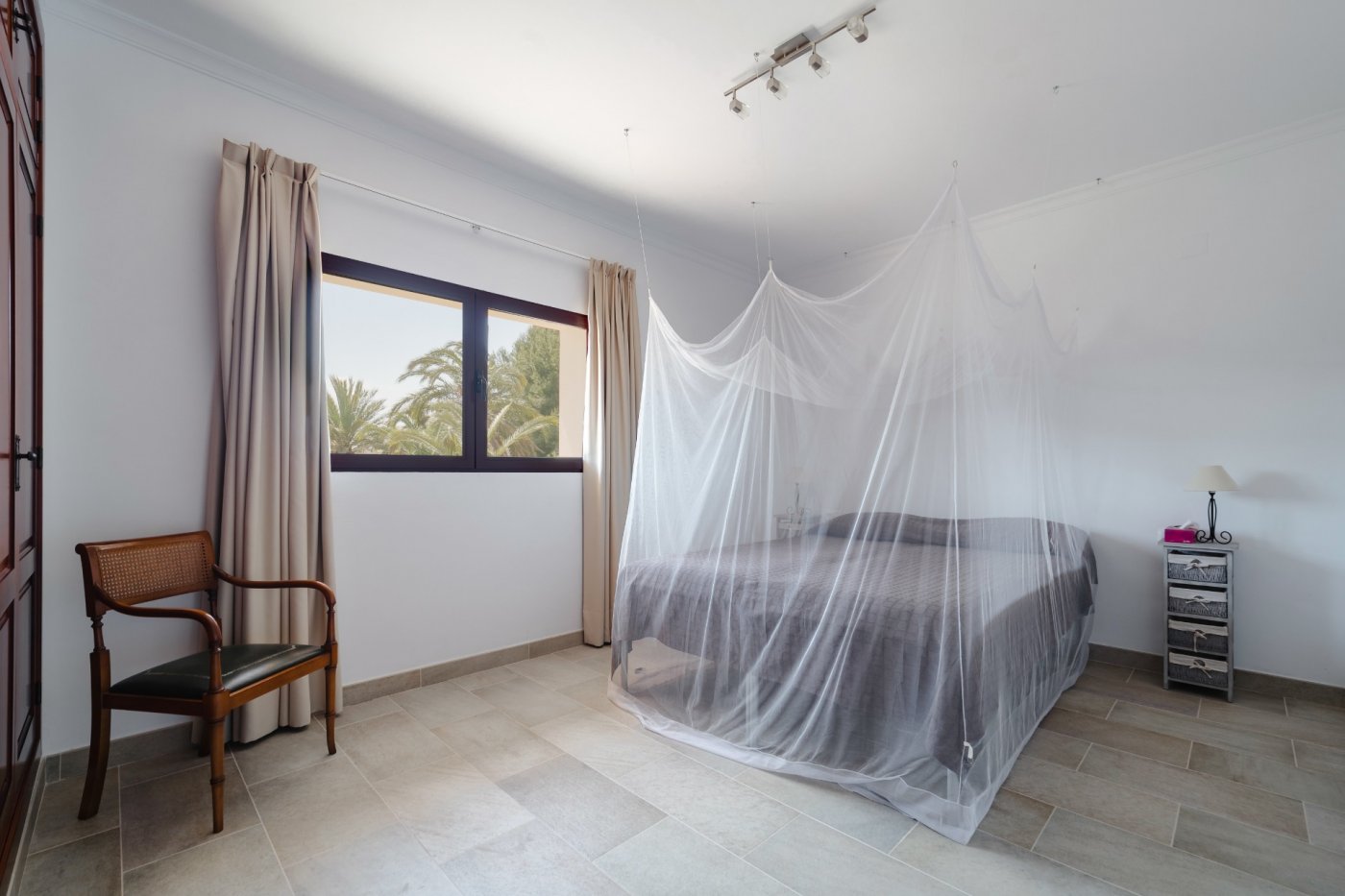 Helles Schlafzimmer mit Doppelbett und Moskitonetz, Keramikfliesenboden und Fenster mit Blick auf den mediterranen Palmengarten.