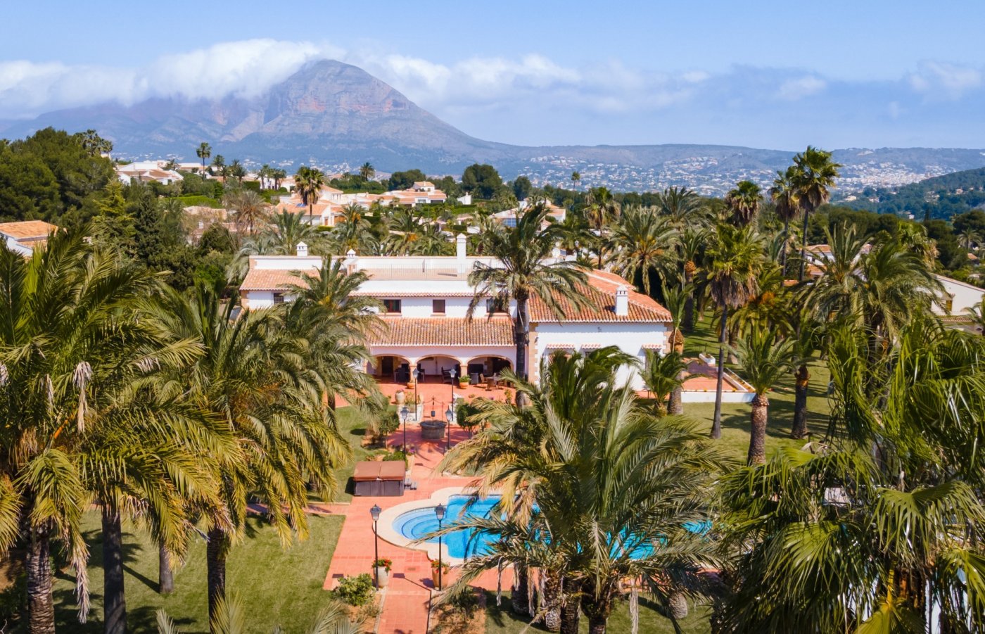 Luftaufnahme der mediterranen Villa mit großem Pool, weitläufigem Palmengarten und Panoramablick auf den Berg Montgó im Hintergrund.