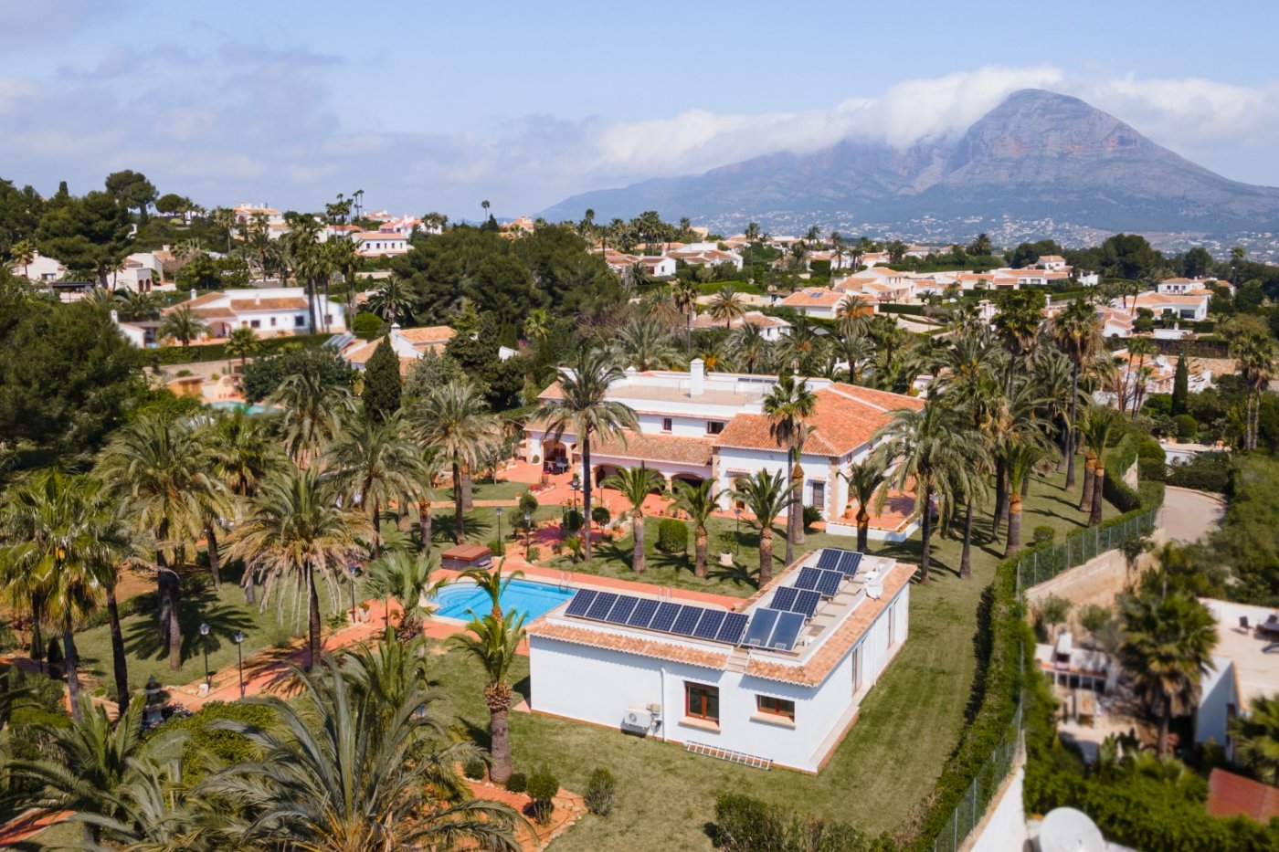 Luftaufnahme des Anwesens mit Haupthaus, Gästehaus mit Photovoltaikanlage, Pool und weitläufigem Palmengarten vor der Kulisse des Montgó-Massivs.