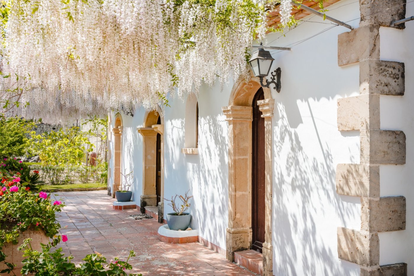 Mediterrane Fassade mit Rundbögen aus Tosca-Stein und Holztüren, beschattet von einer blühenden weißen Blauregen-Pergola auf Terrakottaboden.