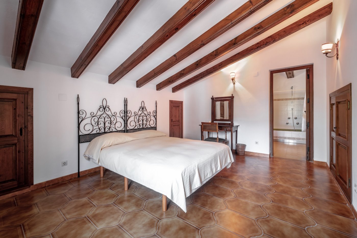 Mediterranes Schlafzimmer mit Dachschräge und Holzbalkendecke, rustikalem Fliesenboden, schmiedeeisernem Kopfteil und direktem Zugang zum En-Suite-Badezimmer.
