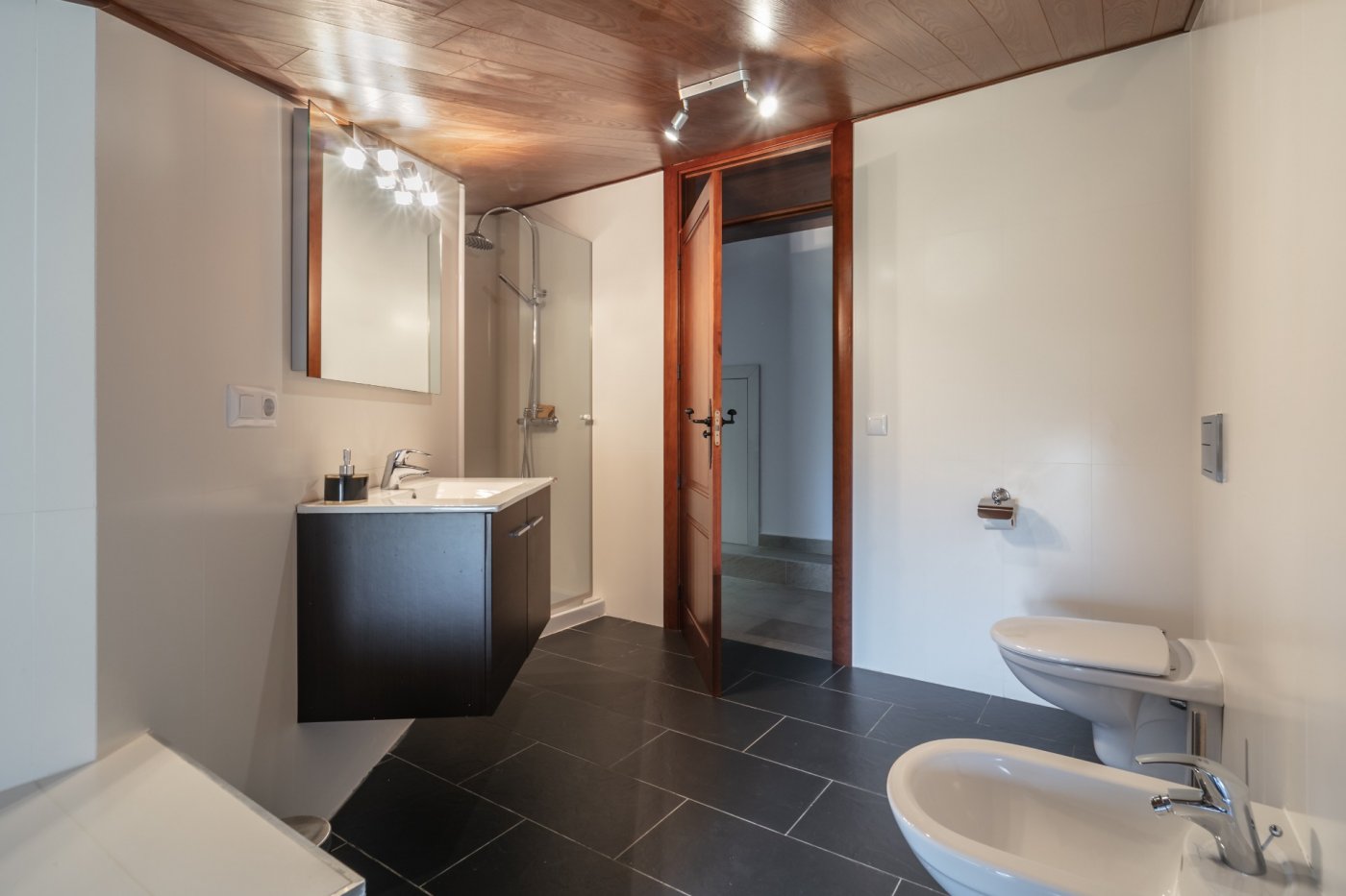 Modernes Badezimmer mit Holzdecke, dunklem Schieferboden, schwebendem Waschtisch, Glasduschkabine und weißen Sanitärobjekten.