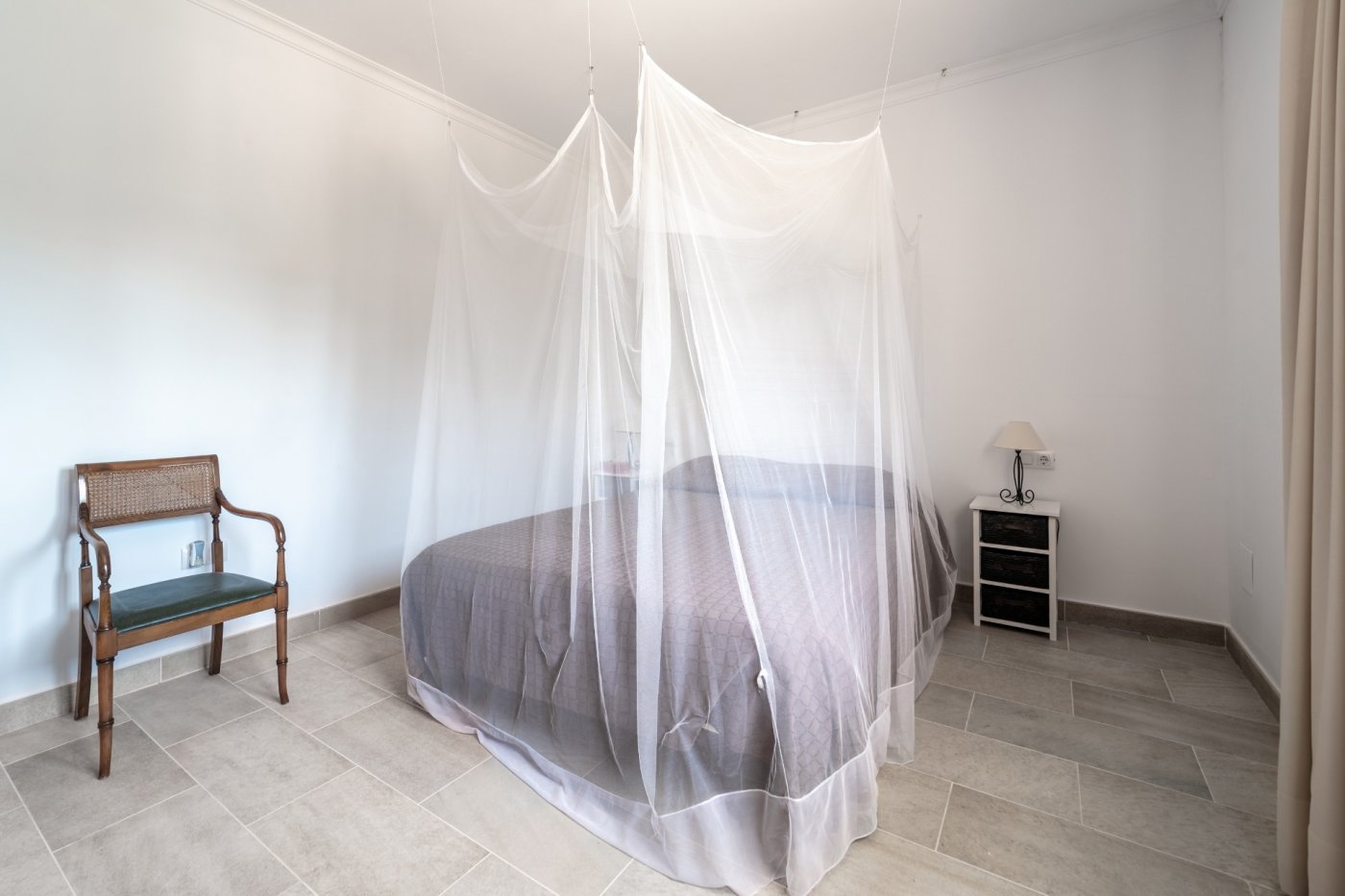 Schlafzimmer mit Doppelbett und großem Moskitonetz, Keramikfliesenboden und einem klassischen Holzstuhl.