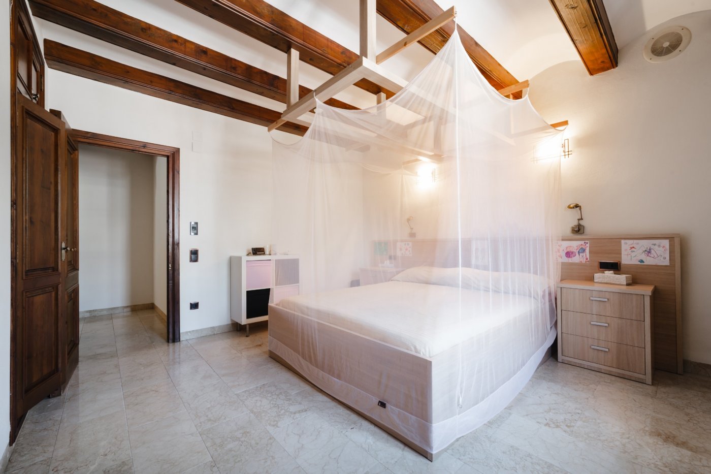 Schlafzimmer mit dunklen Deckenbalken, Bett mit Moskitonetz-Baldachin und hellem Steinboden. Massive Holztüren und integrierte Wandleuchten runden das Bild ab.