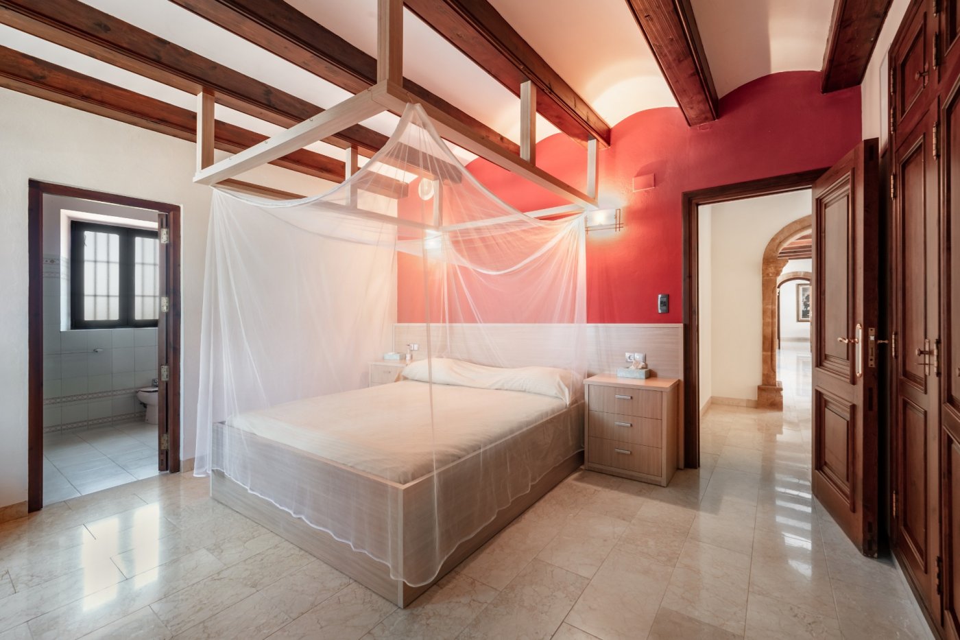 Schlafzimmer mit Holzbalkendecke, Himmelbett mit Moskitonetz, roter Wand und Marmorboden, mit Blick auf das angrenzende Bad und einen Steinfundbogen.