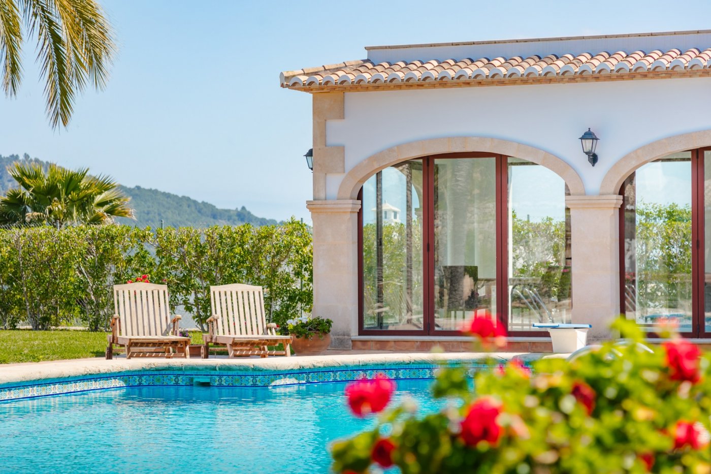 Swimmingpool mit blauen Mosaikfliesen neben einem mediterranen Gebäude mit Tosca-Steinbögen, Holzfensterrahmen und einer Sonnenterrasse mit Liegestühlen.