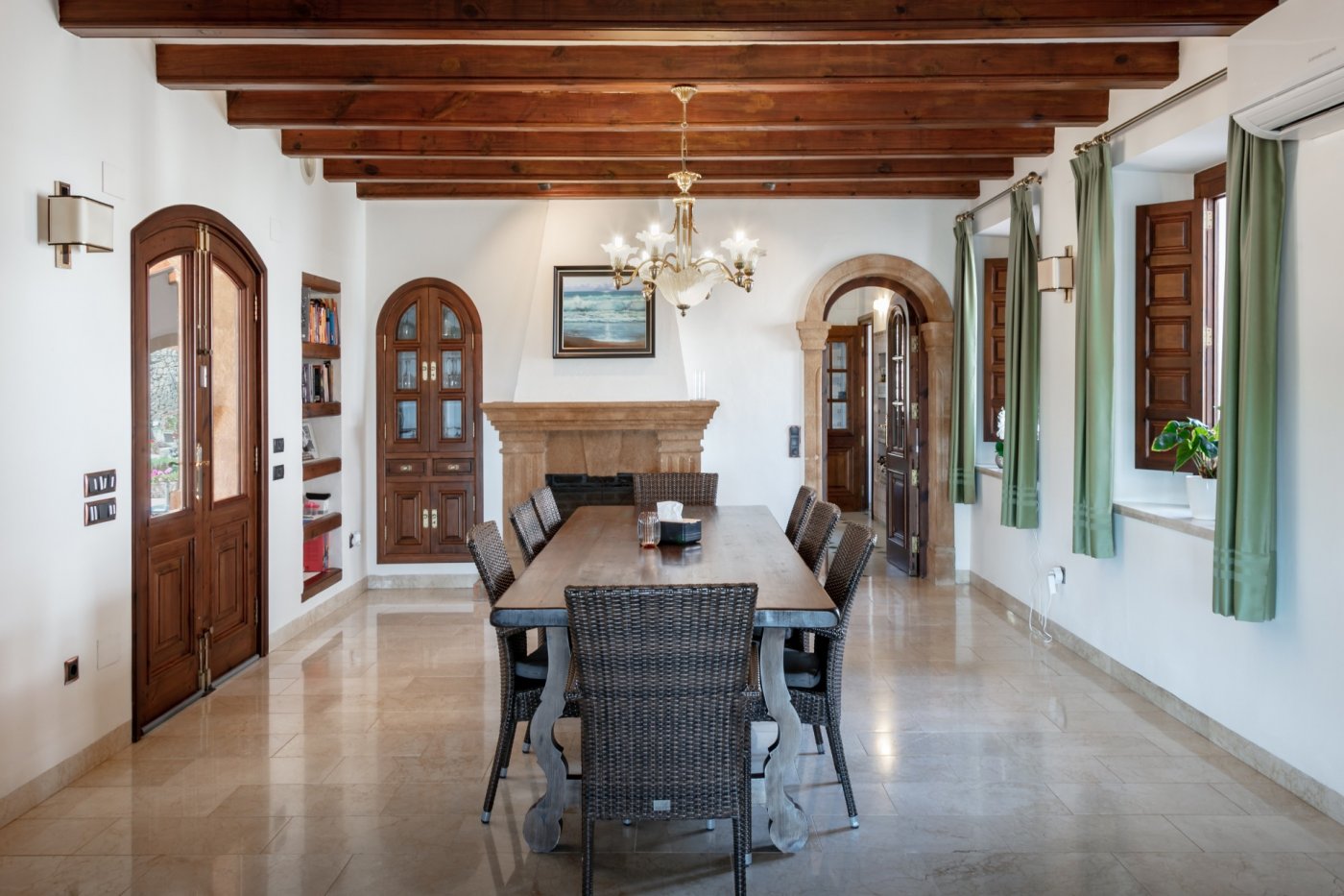 Traditionelles Esszimmer mit sichtbaren Holzbalken, einem Kamin aus Tosca-Stein und Massivholztisch. Steinbögen und Marmorböden unterstreichen den mediterranen Stil.