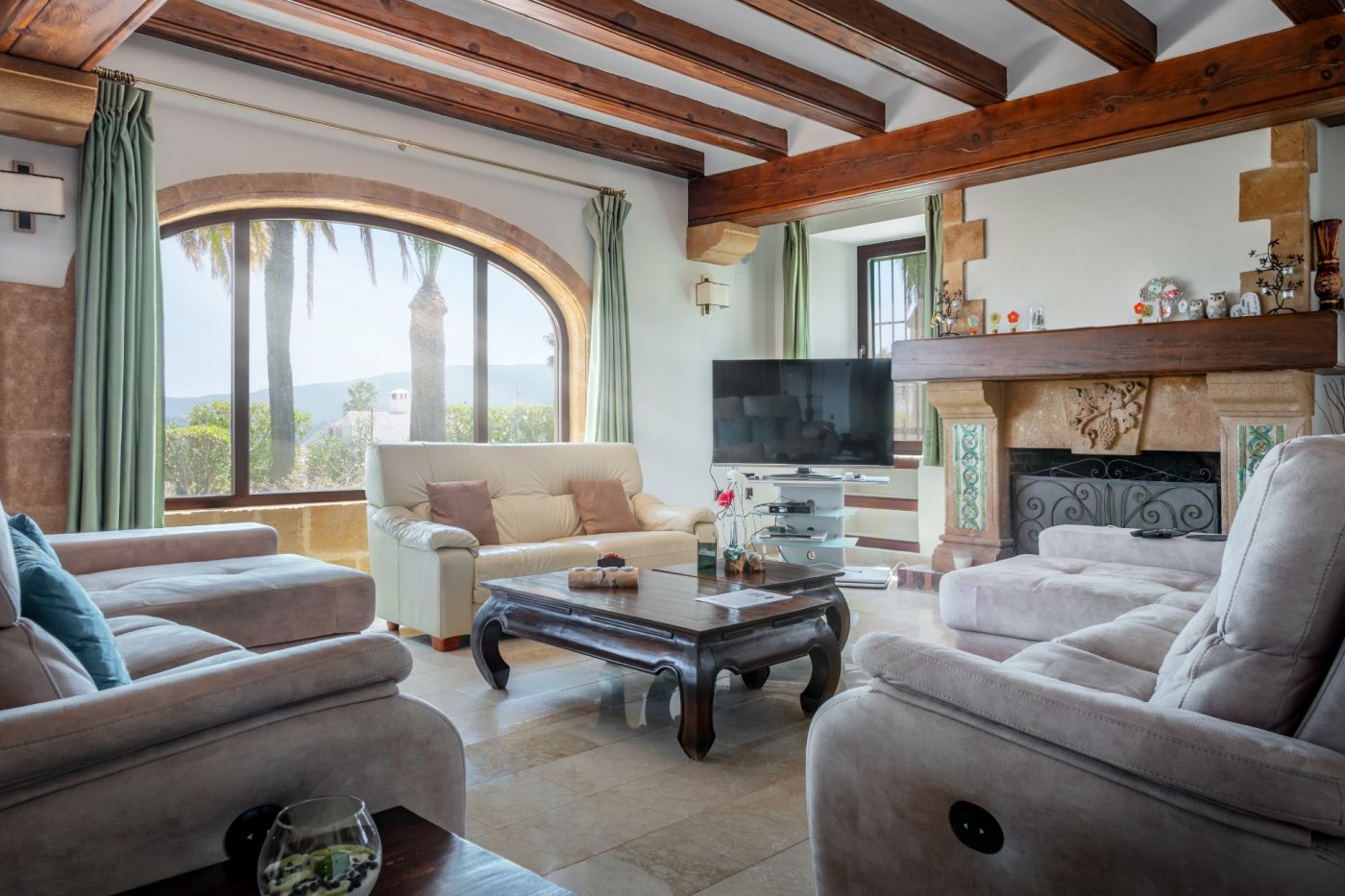 Wohnzimmer im mediterranen Stil mit Holzbalkendecke, großem Rundbogenfenster aus Tosca-Stein und rustikalem Kamin mit Blick auf das Tal.