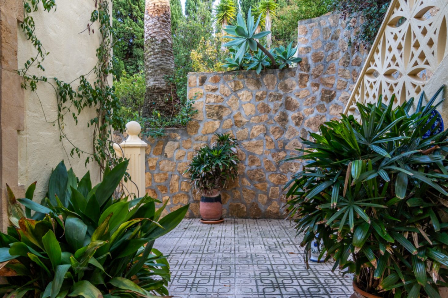 Außenhof mit geometrisch gemusterten Fliesen, Natursteinmauern und üppiger mediterraner Bepflanzung, einschließlich Palmen und Sukkulenten.